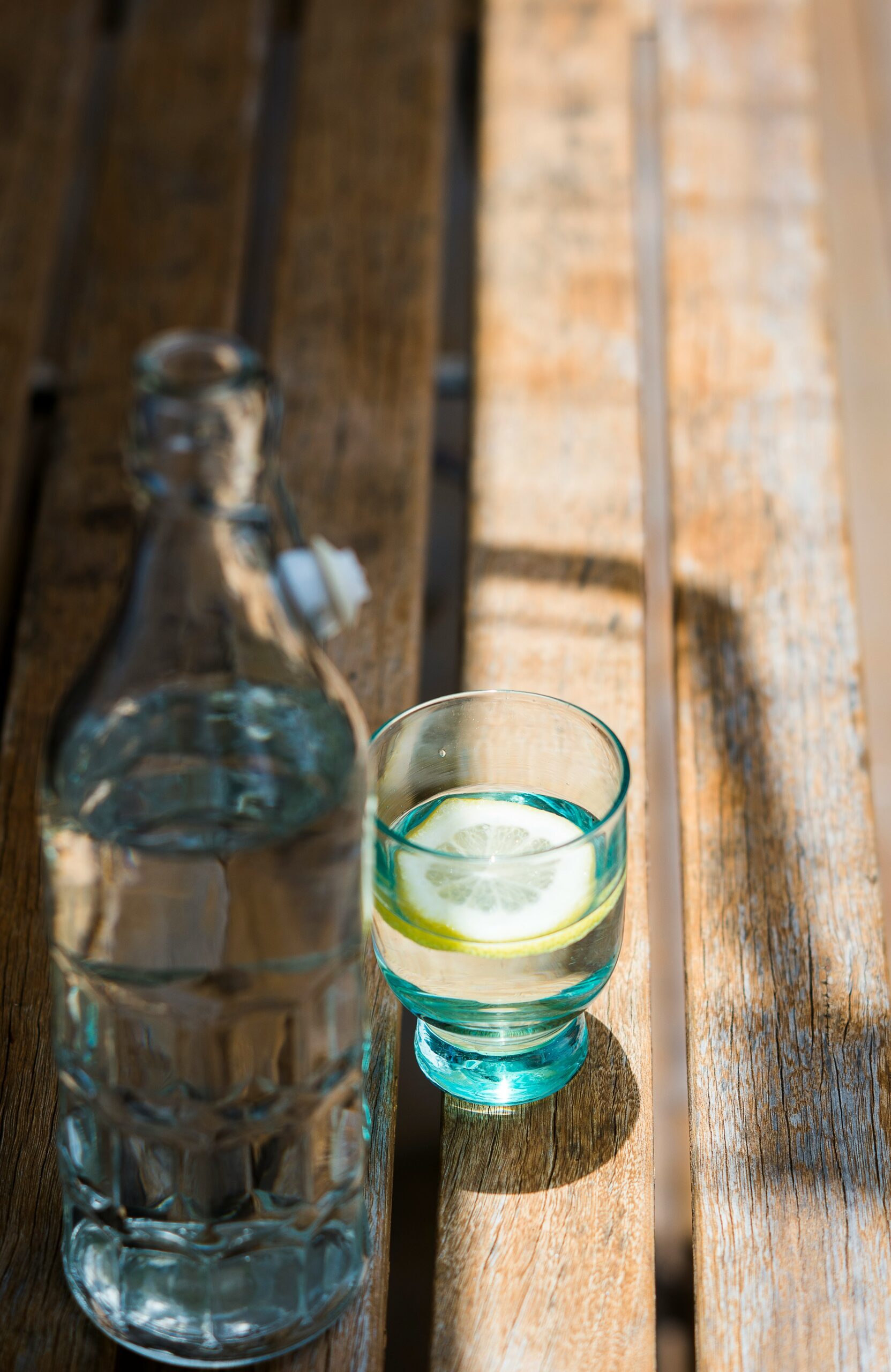 A glass bottle of water and a glass with a lemon slice sit on a wooden table in sunlight.