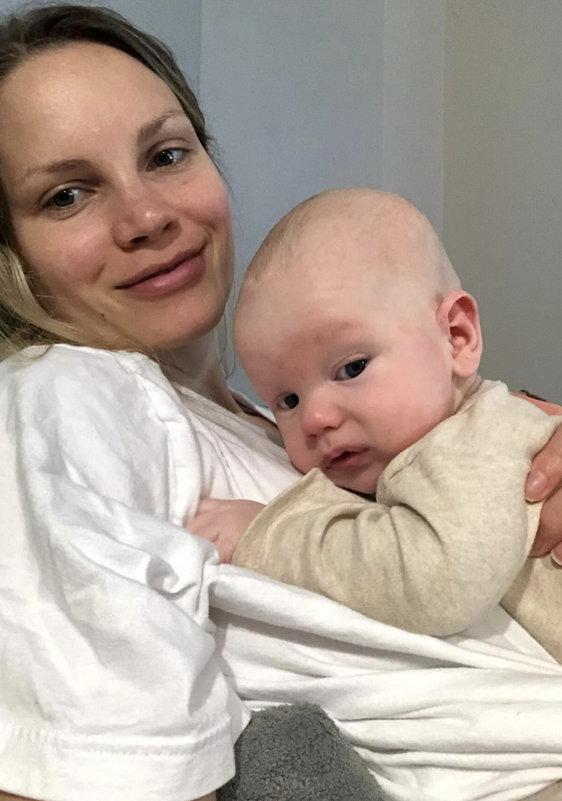 A woman holding a baby while smiling at the camera. The baby is wearing a beige outfit and leaning against the woman.