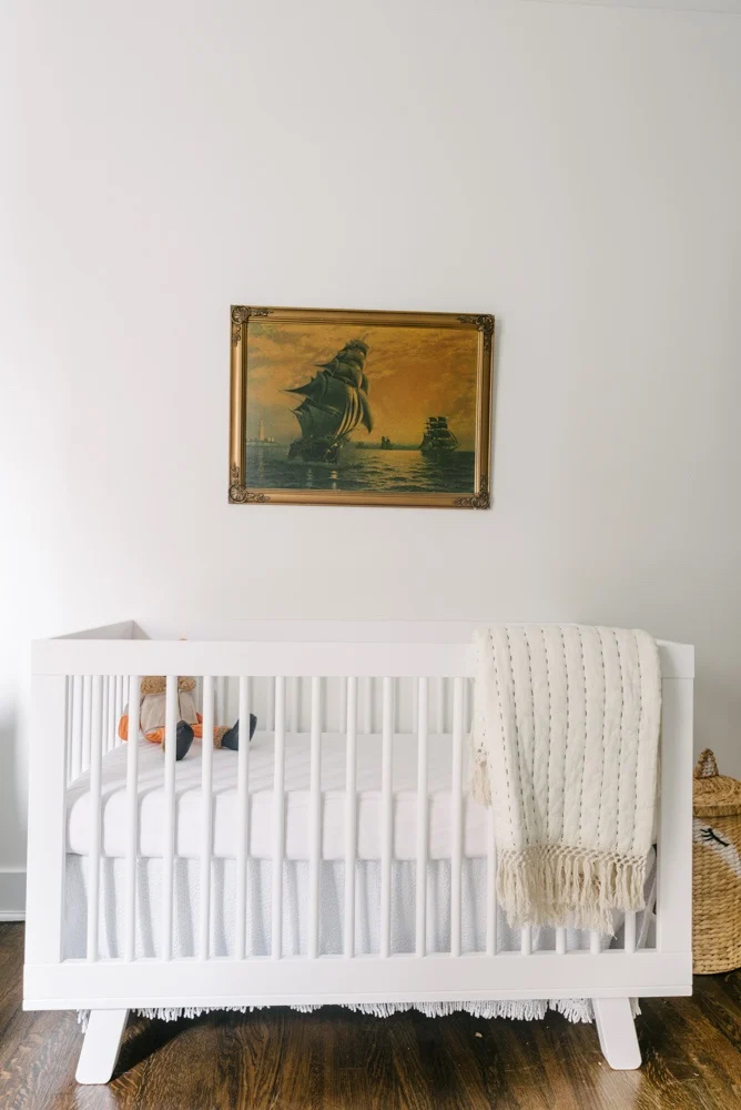 A white crib with a blanket draped over the side stands against a wall. Above it, a framed painting of a ship is hung. A stuffed toy is inside the crib. A woven basket is placed nearby.