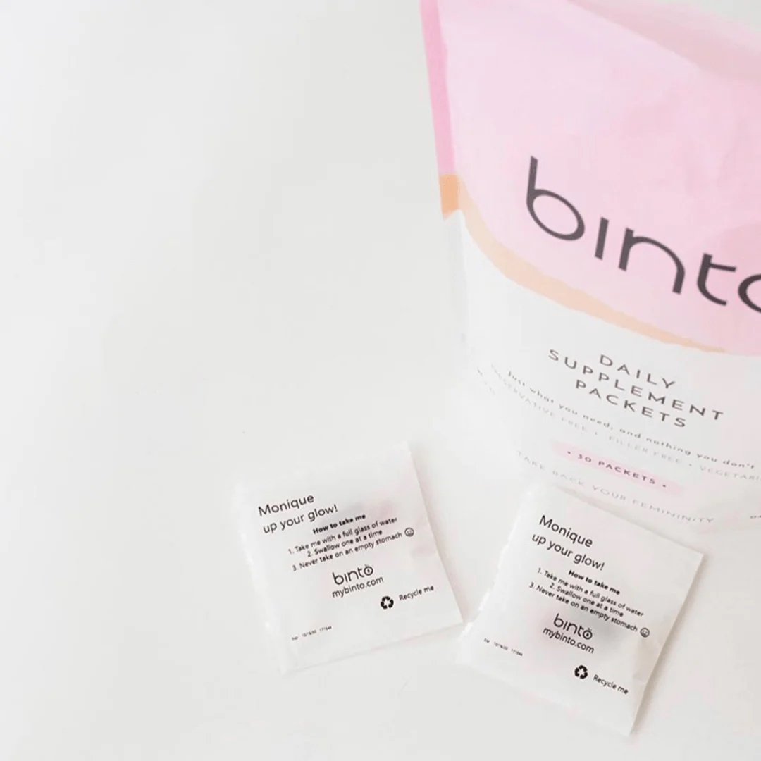 A packet labeled "binto Daily Supplement Packets" sits next to two individual supplement packets that read "Monique up your glow," placed on a white surface.