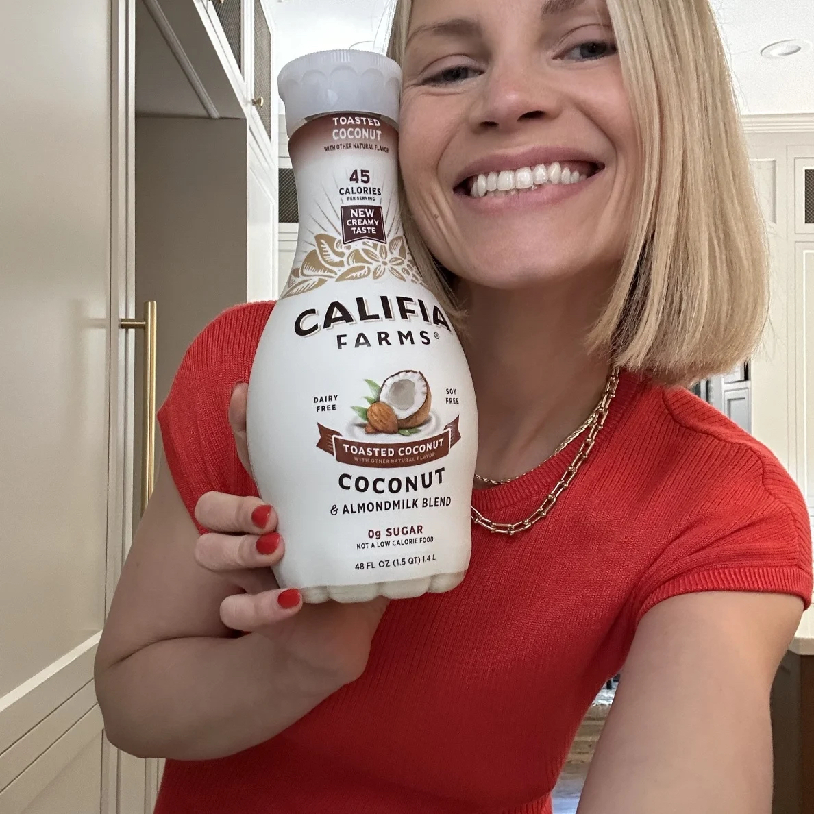 Person in a red shirt smiling and holding a bottle of Califia Farms Coconut & Almondmilk blend in front of a kitchen background.