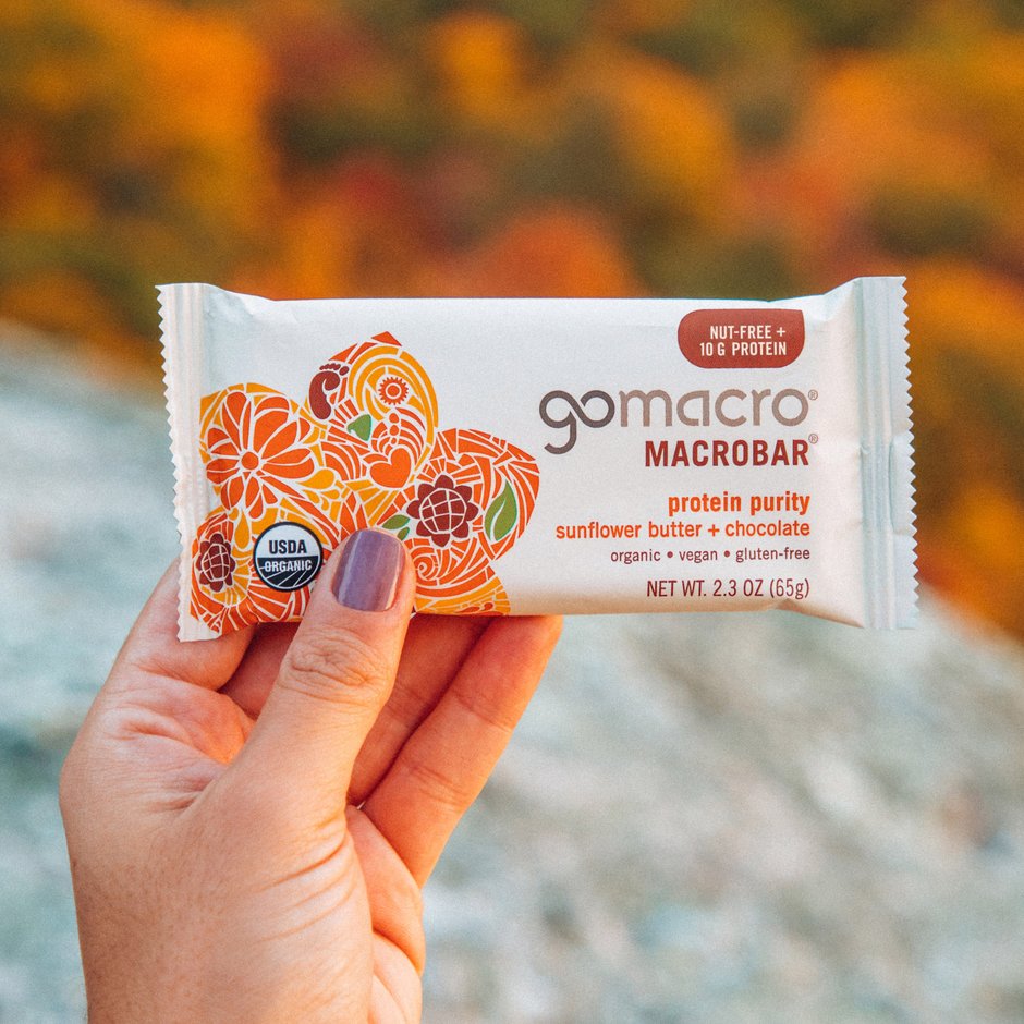 A hand holding a GoMacro MacroBar with protein purity, sunflower butter and chocolate flavor. The packaging indicates it is organic, vegan, gluten-free, and nut-free with 10 grams of protein.