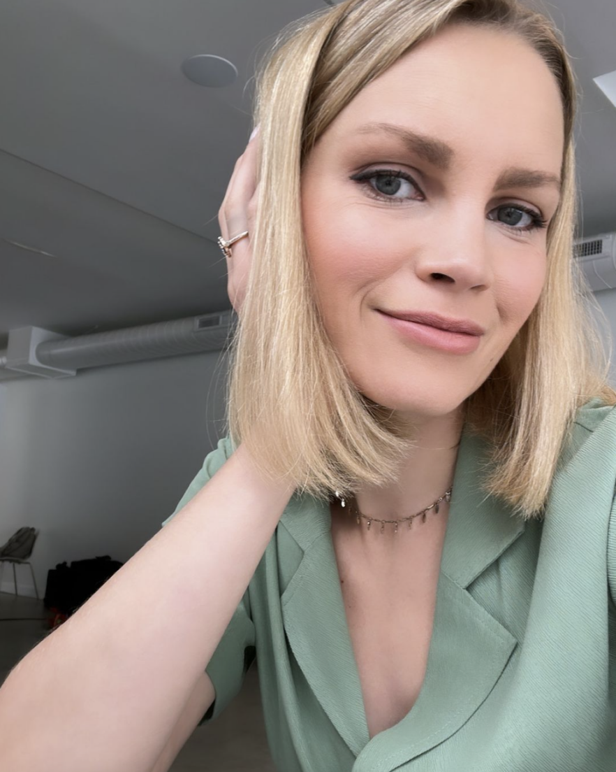 A person with shoulder-length blonde hair and blue eyes, wearing a green top, poses for a selfie indoors.