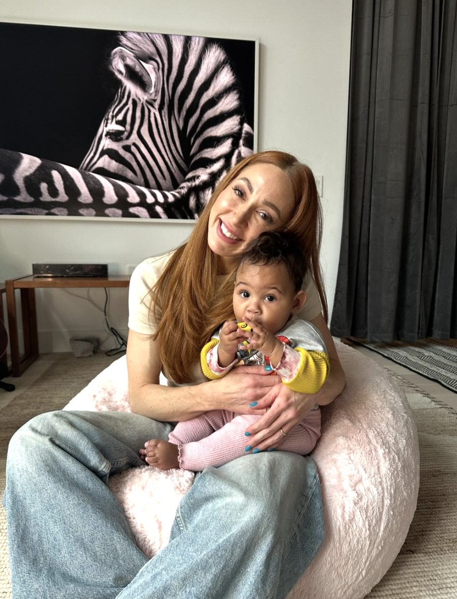 A woman with red hair sits on a beanbag holding a small child with a painting of a Zebra in the background.