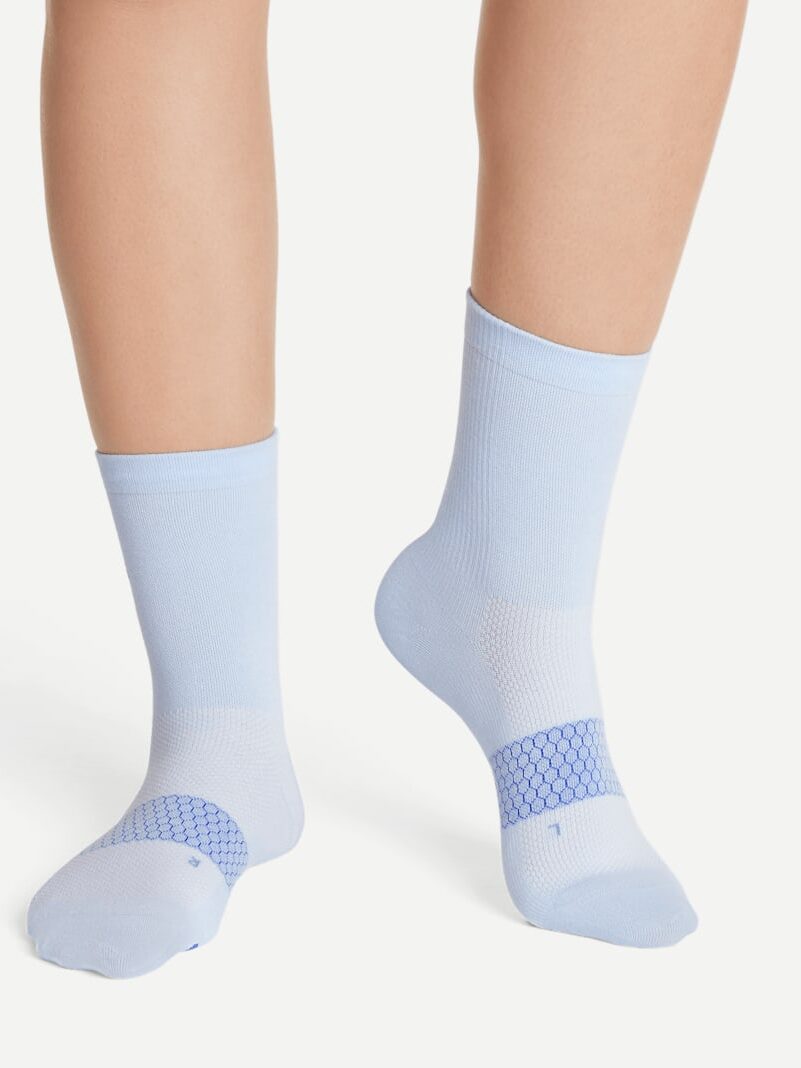 Close-up of a person wearing light blue socks with a darker blue geometric pattern around the arch, against a plain background. Nearby, a sleek massage gun rests, ready to soothe tired feet after a long day.