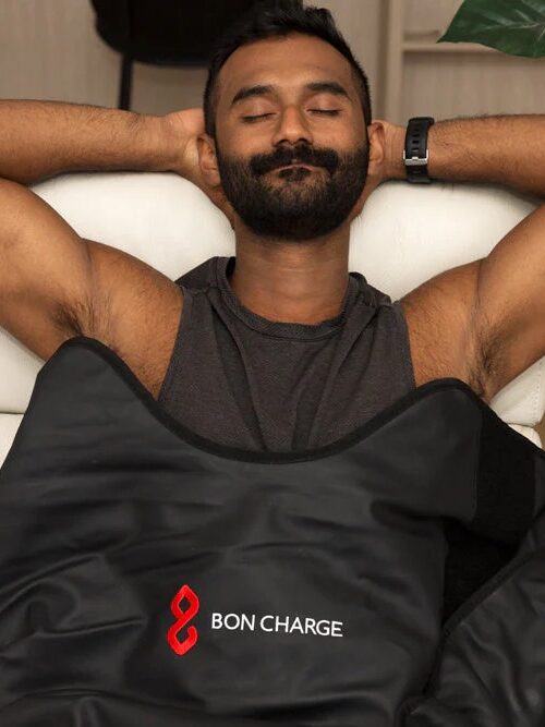 A person unwindingly reclines on a couch with a black BON CHARGE blanket, arms comfortably behind their head. 