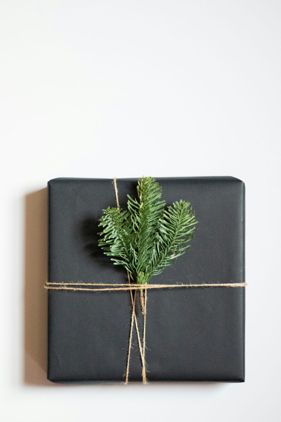 A square gift wrapped in black paper with twine, subtly hinting at the soothing power of massage guns, adorned with small green pine branches against a white background.