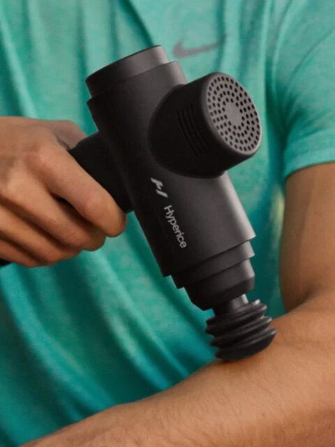 A person in a teal shirt uses a black massage gun on their arm, feeling the soothing relief with each pulse.