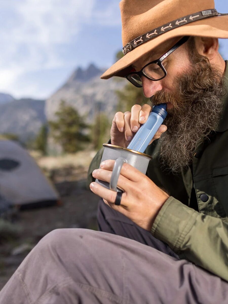 A man uses a portable water filter straw to drink from a metal cup while sitting outdoors, with a tent and mountains in the background. 