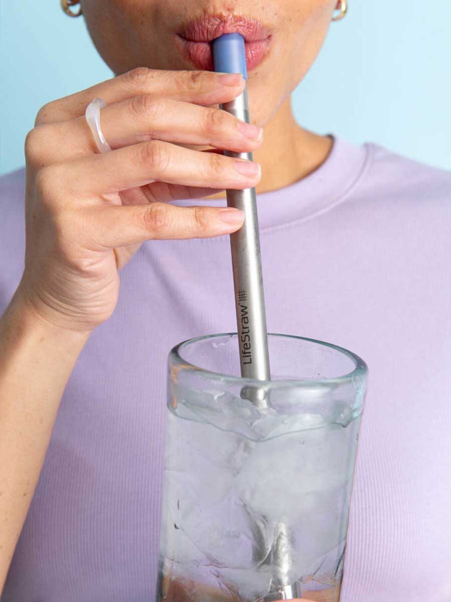 A person drinks from a glass of ice water using a LifeStraw-branded metal straw, wearing a light purple shirt.