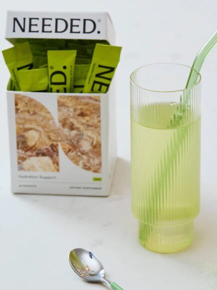 A glass of yellow drink with a straw stands next to a "NEEDED" box on a white surface, alongside a spoon and green packets.