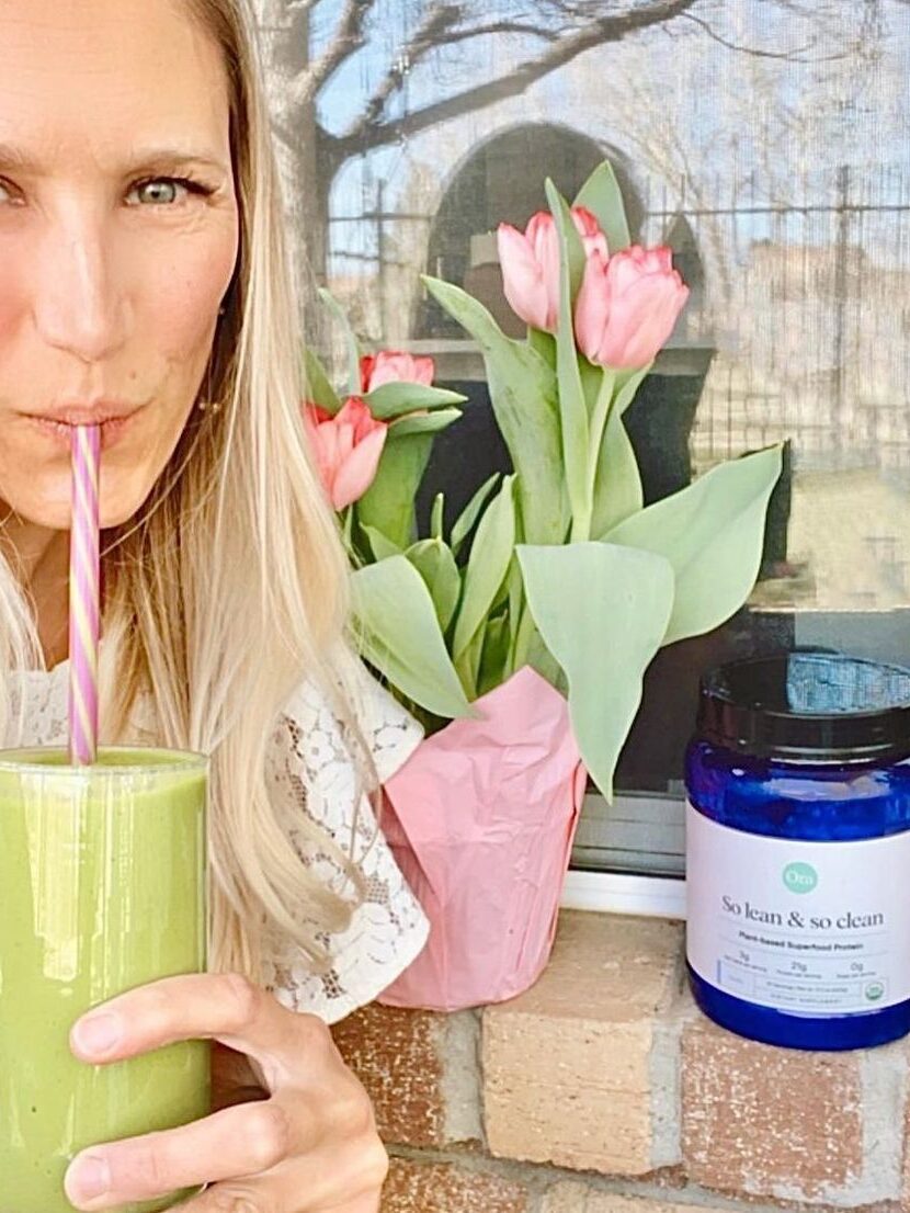 A woman enjoying a green smoothie through a straw sits beside a vase of pink tulips and a blue container labeled "So lean & so clean".