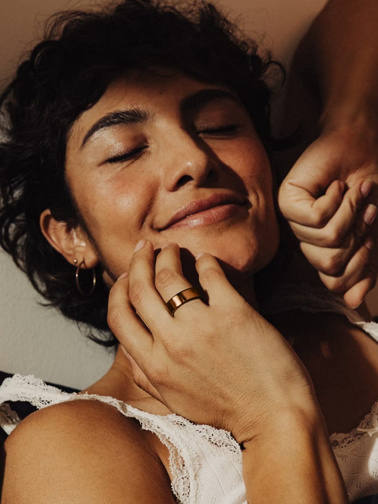 A person with short hair, wearing a white lace-trimmed top and gold ring, smiles softly with eyes closed while touching their face.