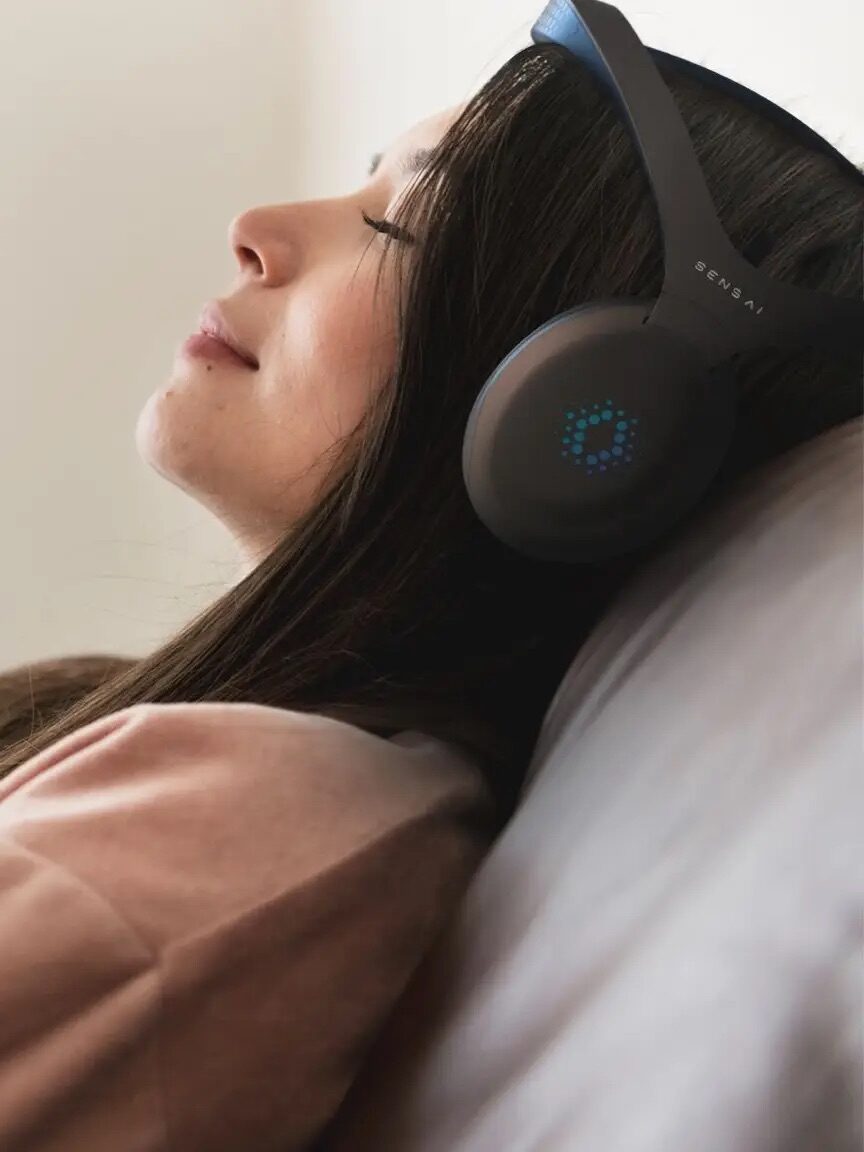 A woman with long brown hair relaxes on a couch, wearing Sens.ai headphones and closing her eyes.