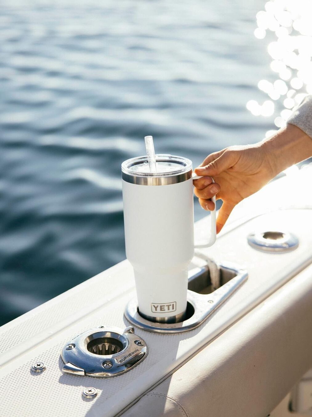 A hand holding a white Yeti tumbler with a straw on a boat near the water.