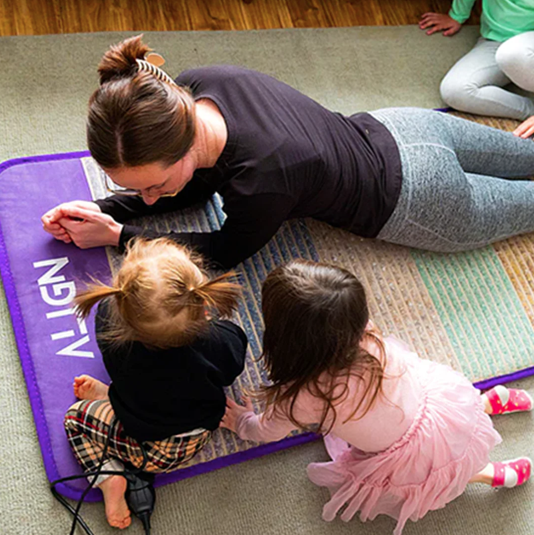 A woman and two children relax on a purple mat, casually engaged with each other. Amidst the wood floor and carpet surroundings, a massage gun sits nearby, adding to their peaceful setting.