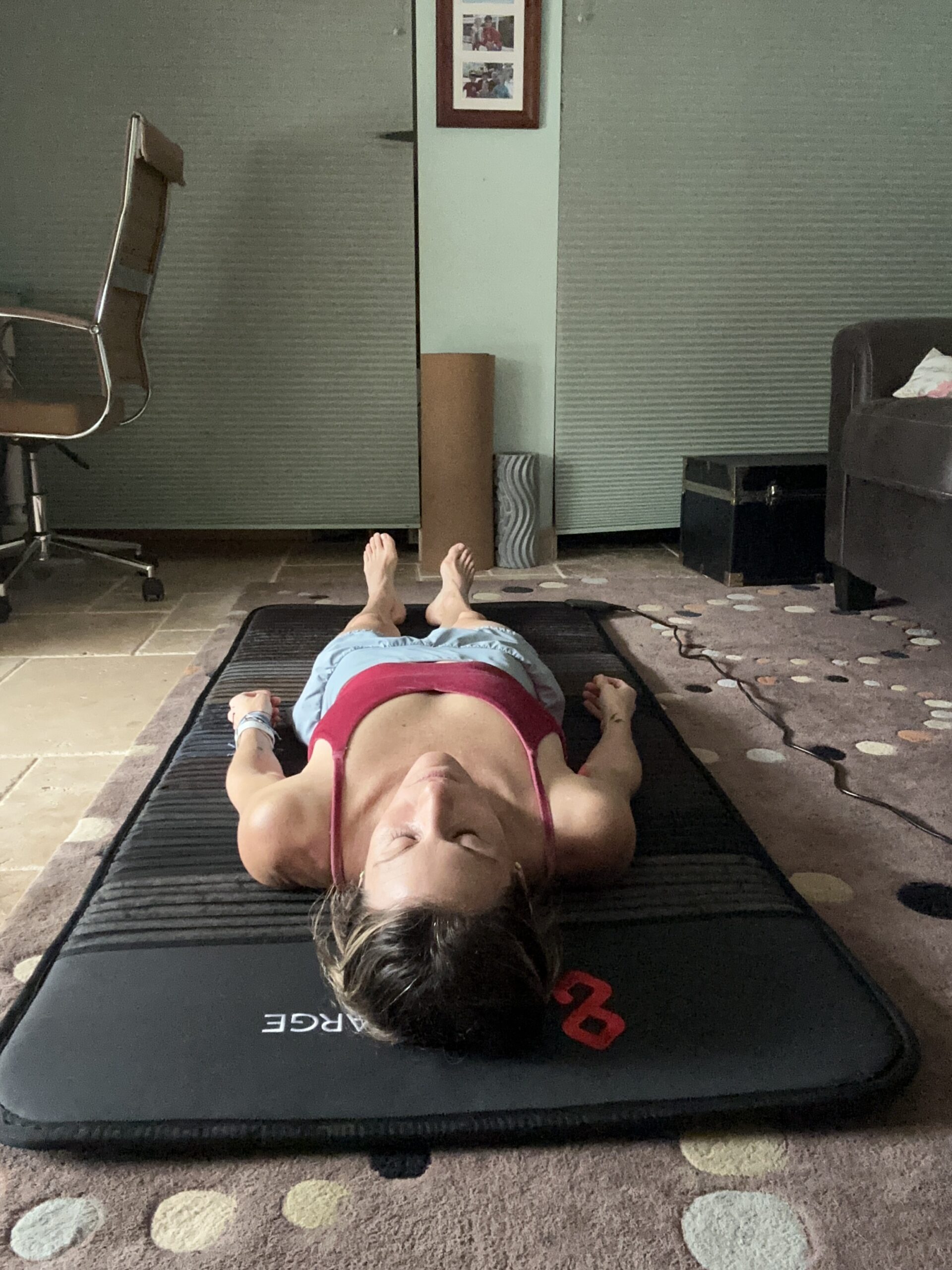 A person in a red top and blue shorts lies on a yoga mat indoors, eyes closed and arms at their sides, as an office chair and furniture surround them. Nearby, a massage gun rests within reach, offering another avenue for relaxation.