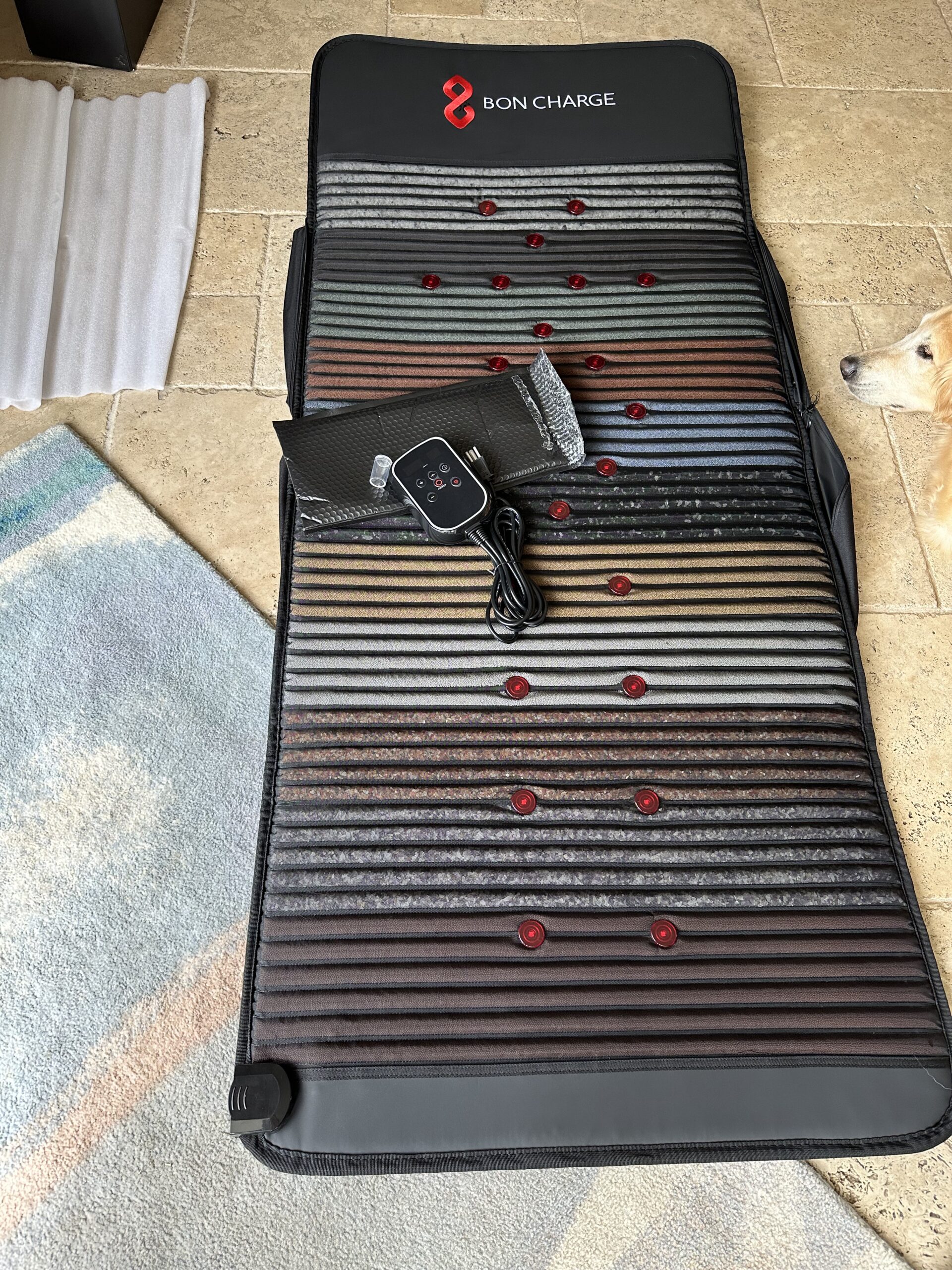 A striped massage mat with red massage nodes rests on a tile floor, accompanied by a control panel and attached wiring. Alongside, massage guns await their turn. A rug and part of a dog's curious face peek into view.