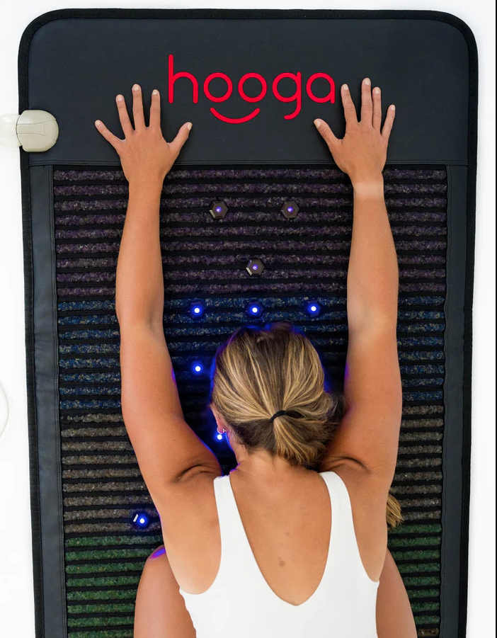 A person lies face down on a mat with outstretched arms, wearing a white top. The "hooga" branded mat bathes them in soothing blue lights, offering a serene experience as massage guns hum gently in the background.