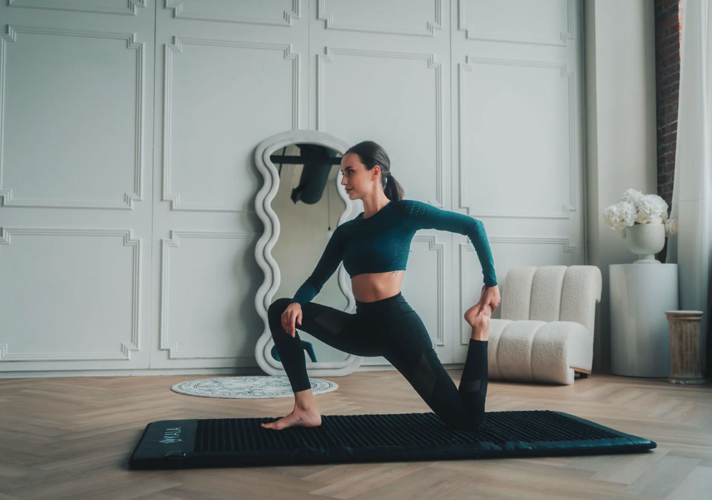 A person in workout attire uses a massage gun while performing a stretching exercise on a black mat in a stylish room with a large wavy mirror and elegant decor.