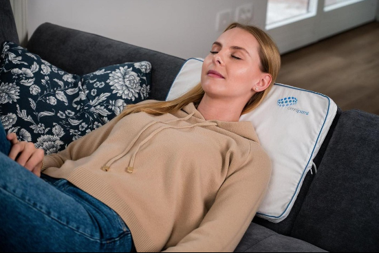 A person laying on a couch with eyes closed, resting on a floral pillow and a white cushion, while a massage gun rests nearby, promising relaxation at their fingertips.