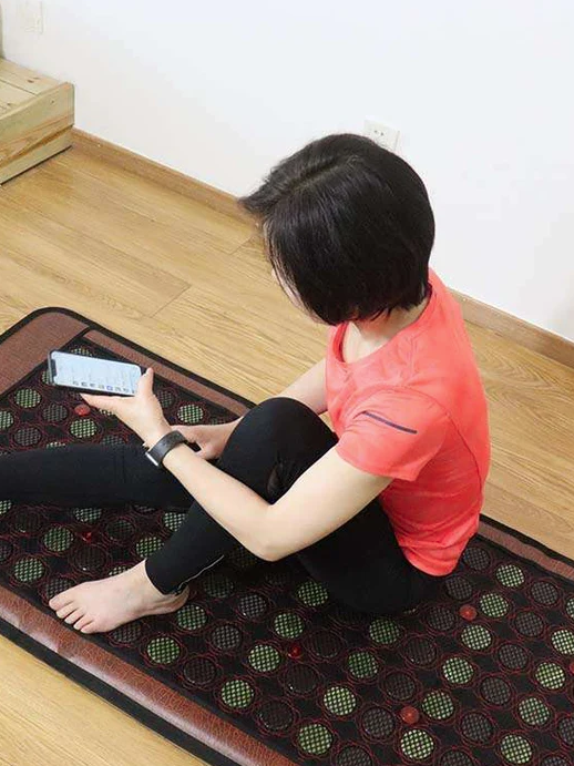 A person relaxes on a heated massage mat, casually scrolling through their phone while contemplating the benefits of massage guns for enhanced muscle relief.