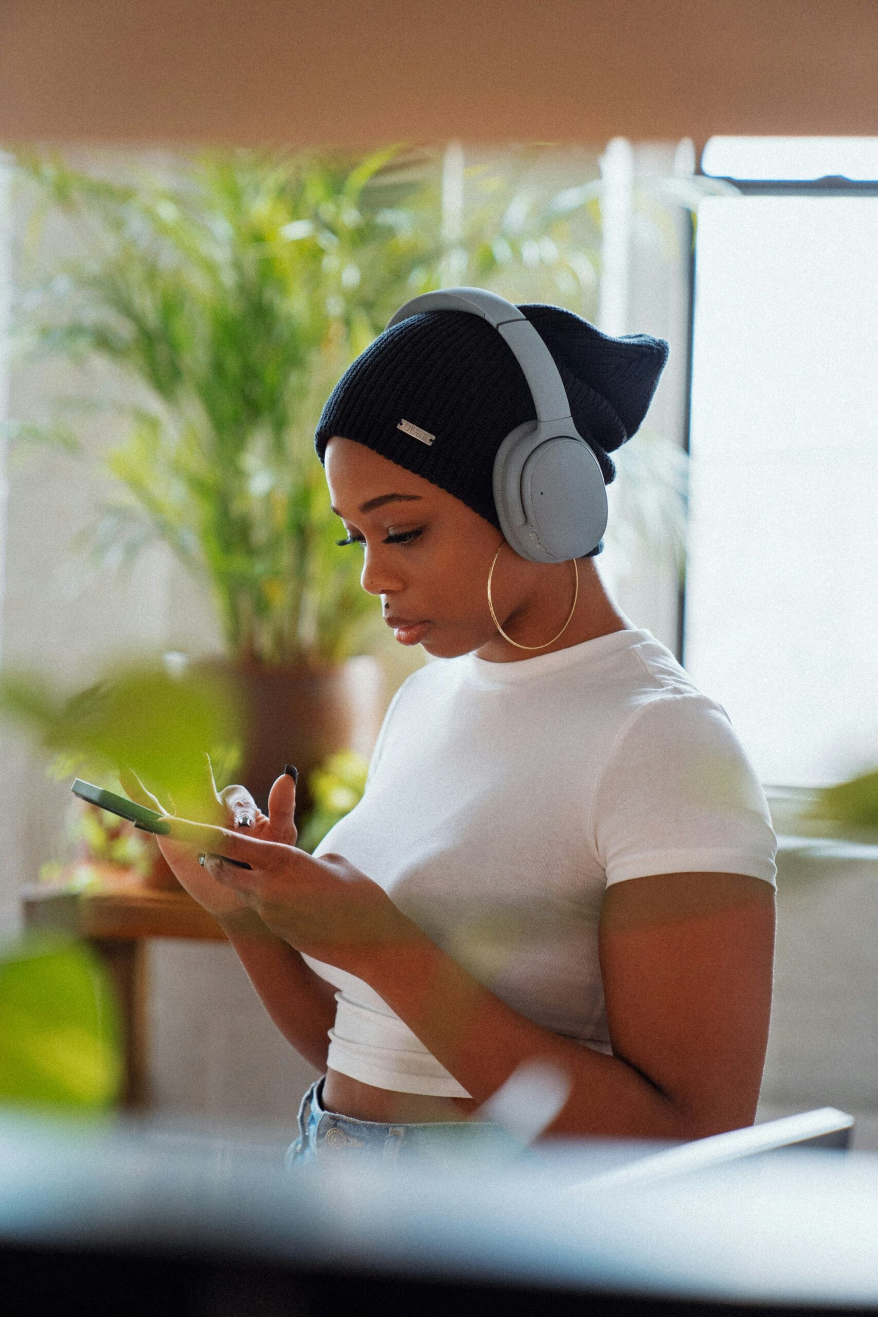 A person wearing headphones and a beanie is looking at a smartphone while standing in a room filled with plants, natural light, and massage guns.