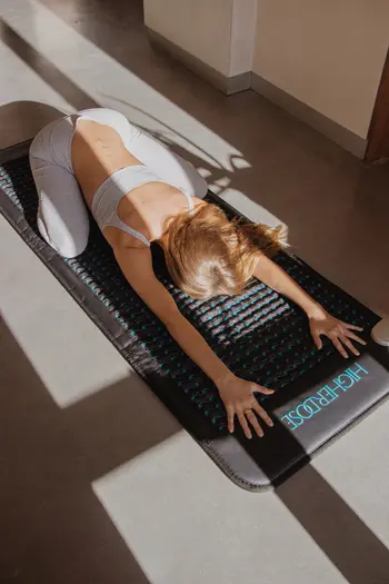 Person in a child’s pose on a yoga mat indoors, with sunlight streaming through a window, contemplating the soothing power of massage guns for post-yoga relaxation.