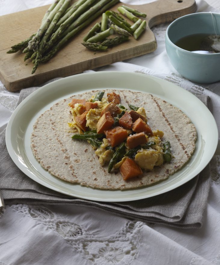 A plate with a tortilla topped with scrambled eggs, sweet potatoes, and asparagus. A cutting board with asparagus and a blue cup are in the background.