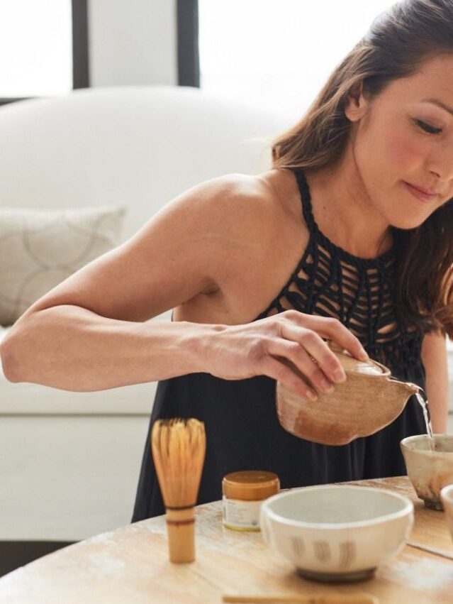 A person pours liquid into a bowl at a table with a whisk and small container, in a bright room with a couch in the background.