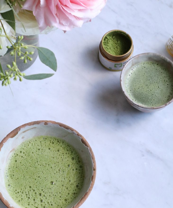 Ceramic bowls with matcha green tea on a marble surface, accompanied by a small container of matcha powder and a bouquet of pink flowers.