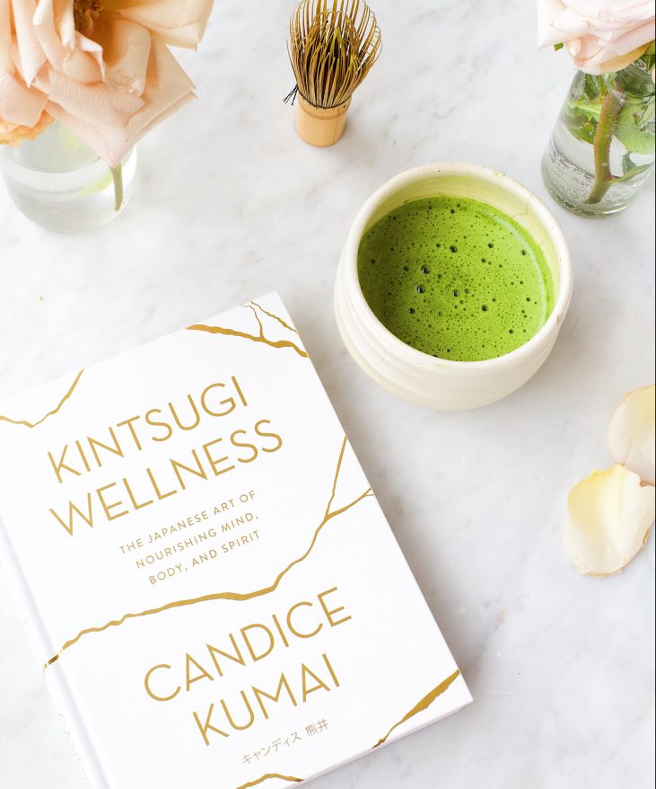 Book titled "Kintsugi Wellness" by Candice Kumai on a marble surface next to a green matcha tea and a small plant.