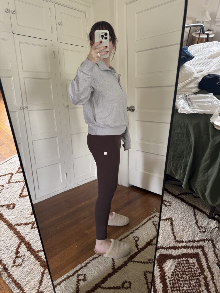 Person in a gray sweatshirt and brown leggings takes a mirror selfie in a bedroom with white cabinets and a patterned rug.