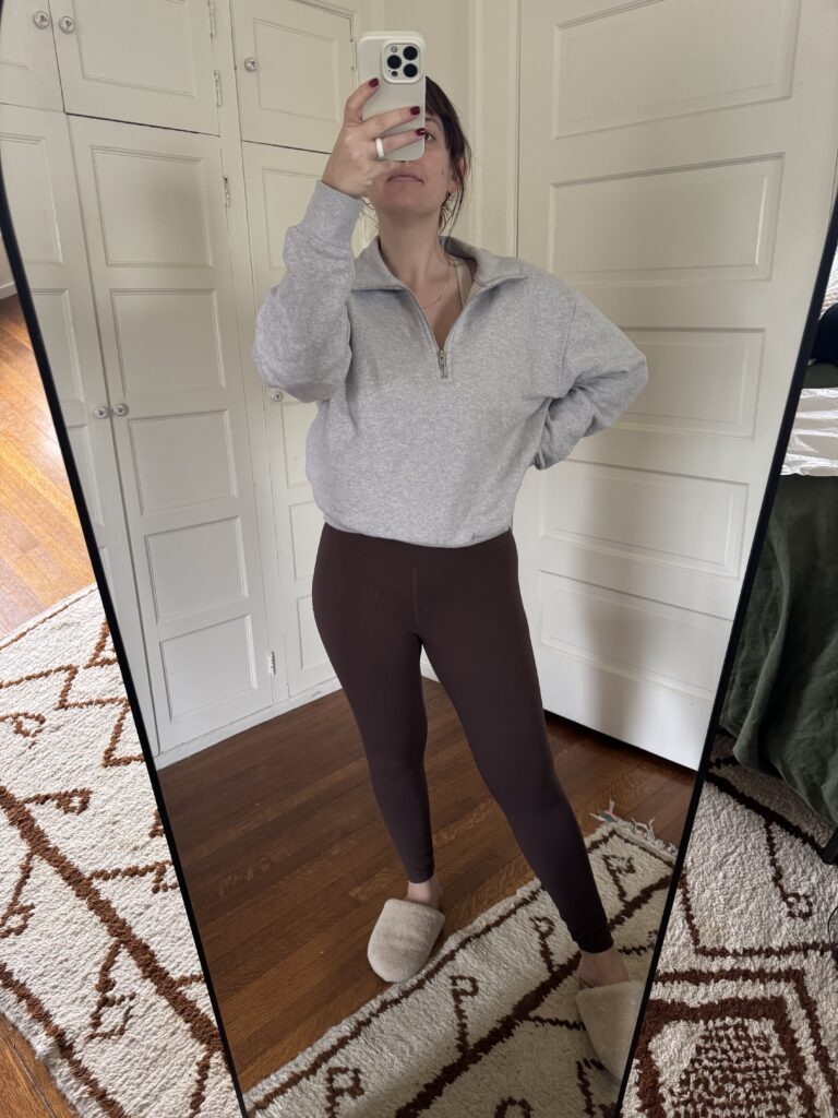 A person wearing a gray pullover, brown leggings, and slippers takes a mirror selfie in a room with wooden floors and a patterned rug.