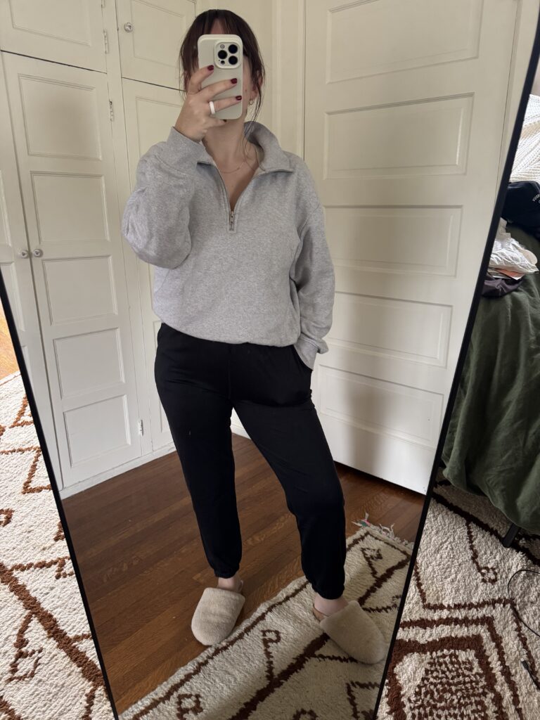 Person wearing a gray pullover, black pants, and slippers taking a mirror selfie in a room with patterned rug.