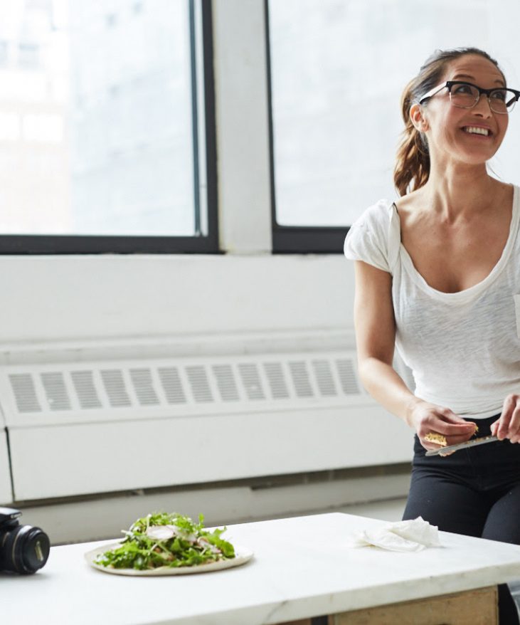 Person with glasses sitting at a table indoors, smiling, with a camera and a plate of salad nearby.