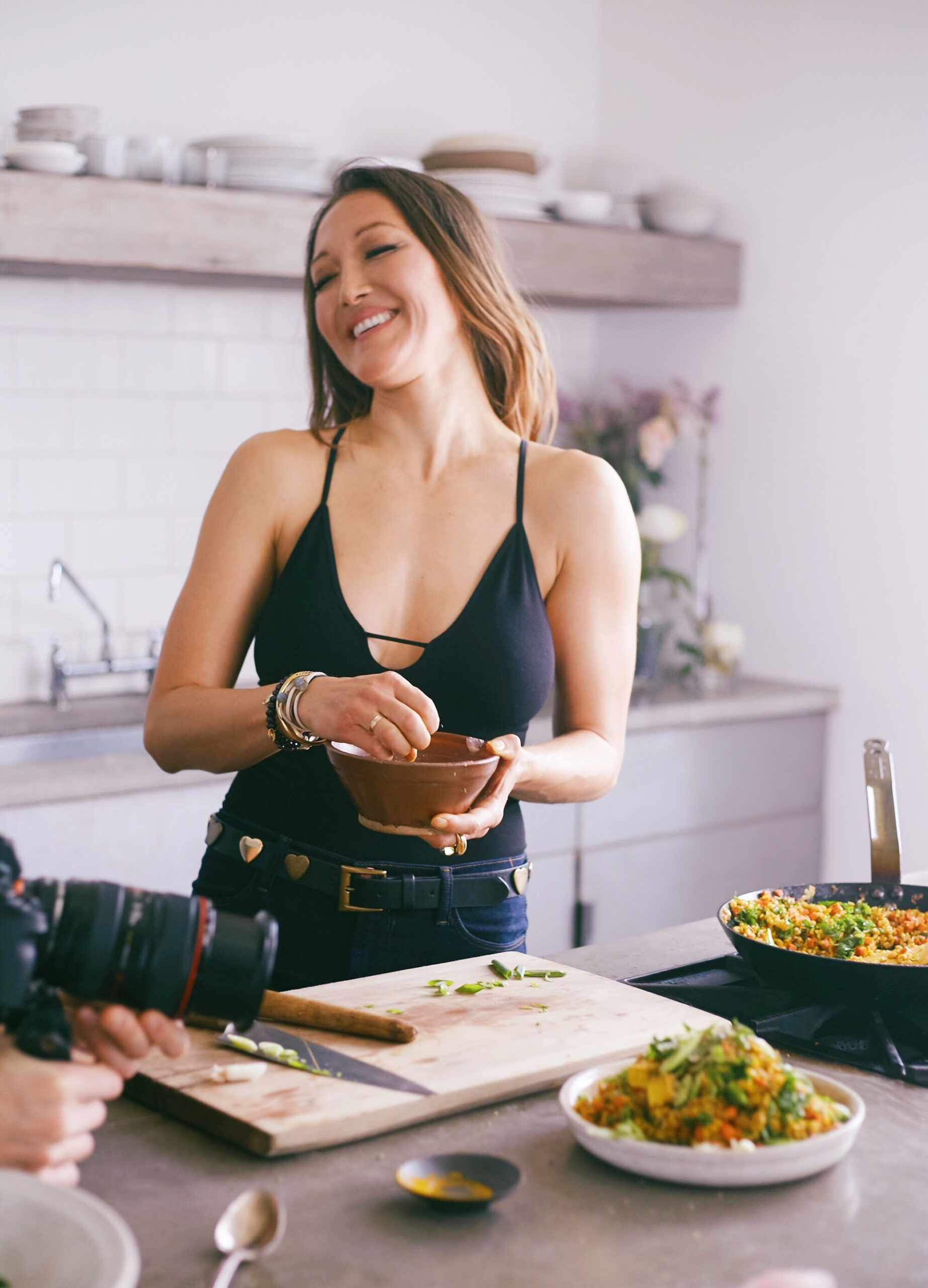 Person in a black tank top smiling while preparing food in a kitchen. A camera is partially visible in the foreground.