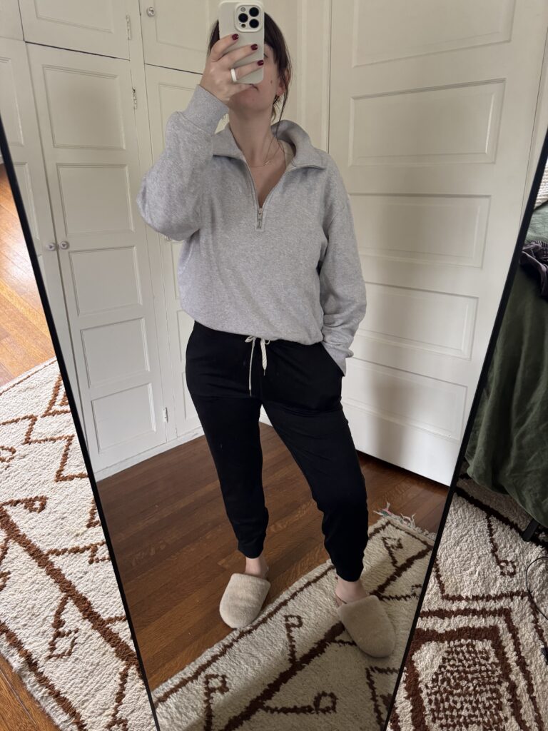 Person taking a mirror selfie wearing a gray sweatshirt, black joggers, and beige slippers. They are in a room with a patterned rug and white doors.