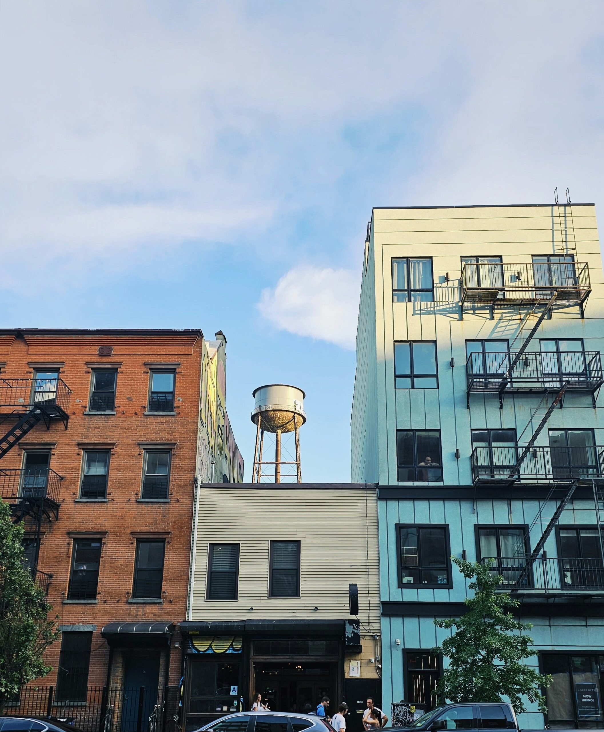 Street view of three adjacent buildings with a water tower in the background. The sky is partly cloudy, and several people are visible near the building entrances.