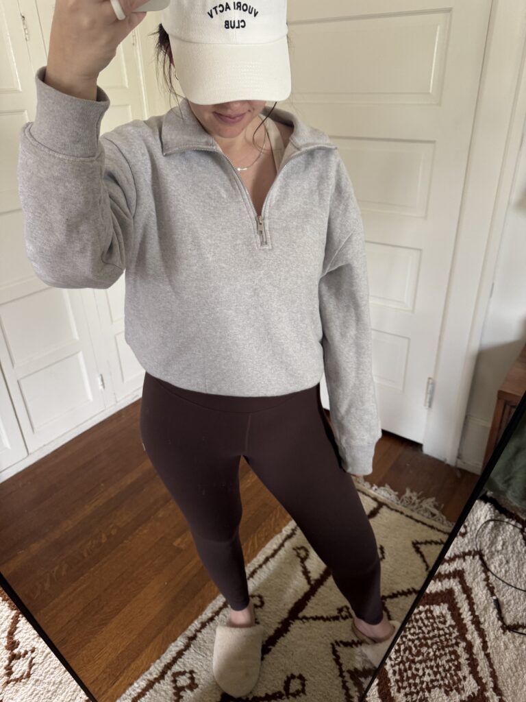 Person in a gray sweatshirt, brown leggings, and a white cap stands on a patterned rug in front of a white door, taking a mirror selfie.