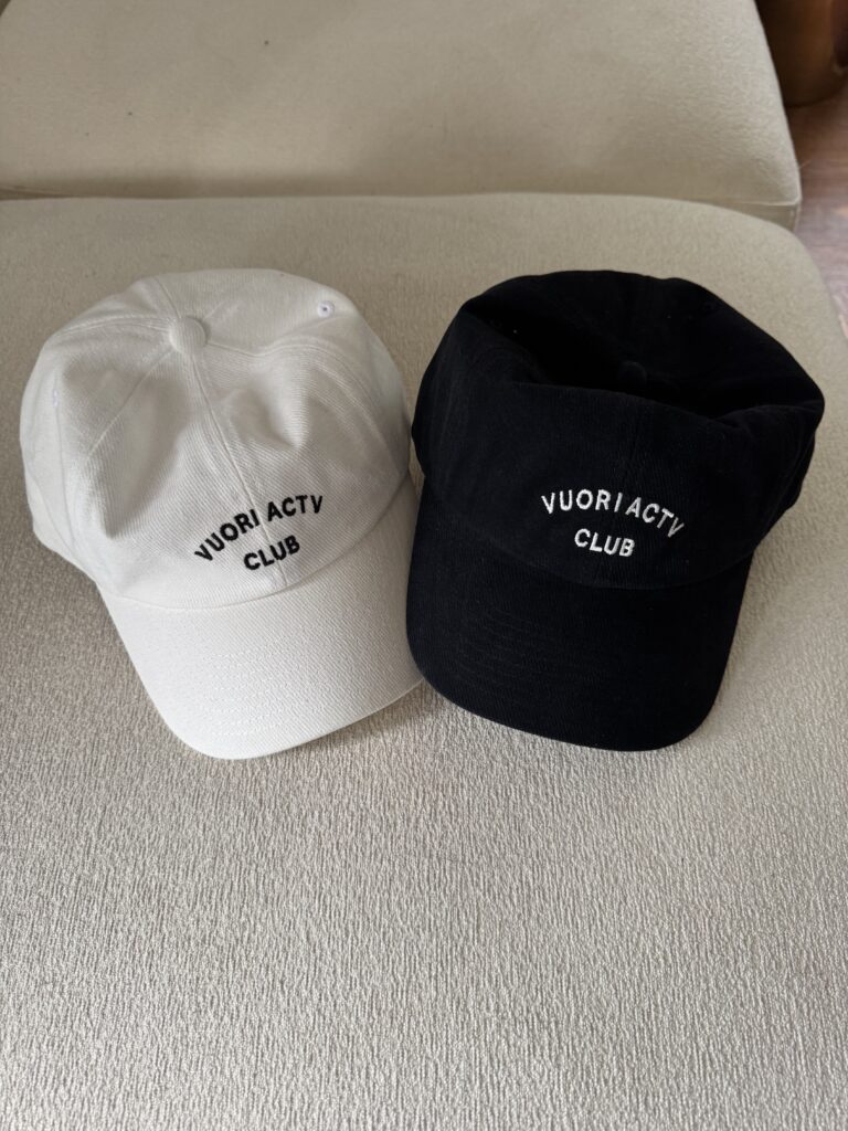 Two baseball caps, one white and one black, both with "VUORI ACTIV CLUB" text on the front, placed side by side on a light-colored surface.