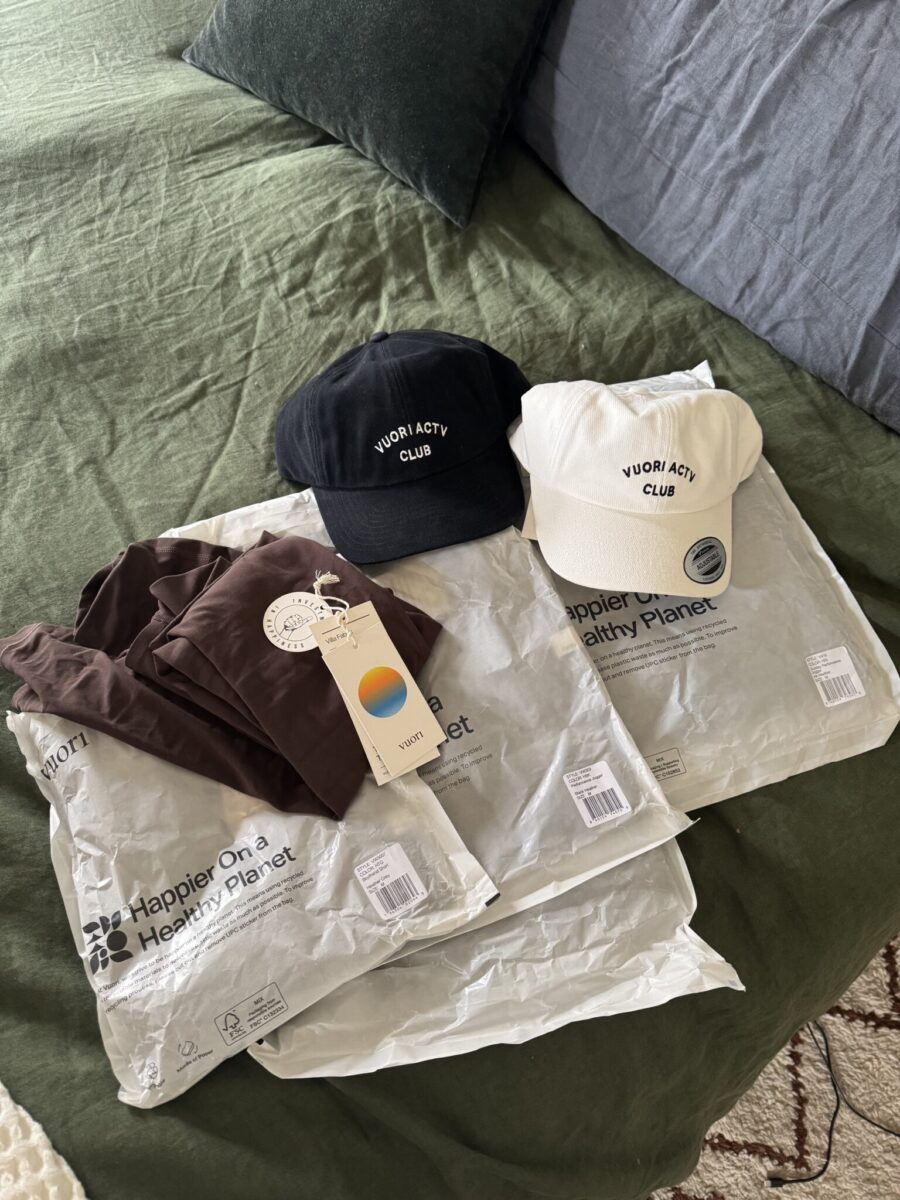 Two "Vuori ACTV Club" caps, one black, one white, placed on a bed with two shirts, packaging, and tags from a brand focusing on eco-friendly initiatives.