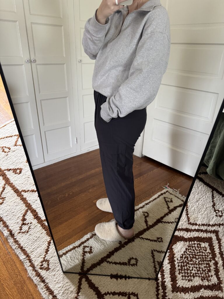 Person wearing a gray sweatshirt, black pants, and slippers stands sideways, taking a mirror selfie in a room with white doors and a patterned rug.