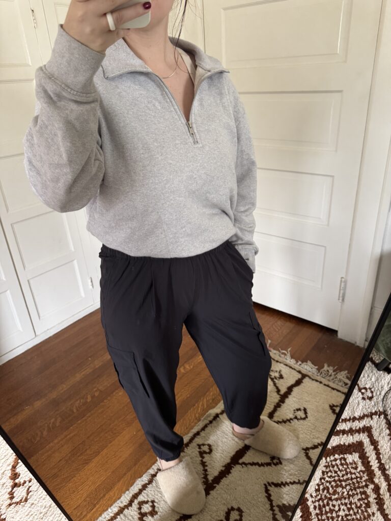 Person in a gray zip-up sweatshirt, black pants, and slippers taking a mirror selfie.