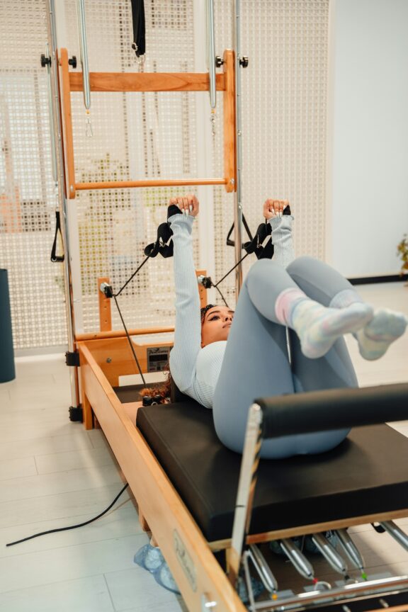 In a well-lit studio, a person exercises on a Pilates reformer machine, wearing a long-sleeve top and leggings. Nearby, massage guns are neatly arranged for post-workout recovery.