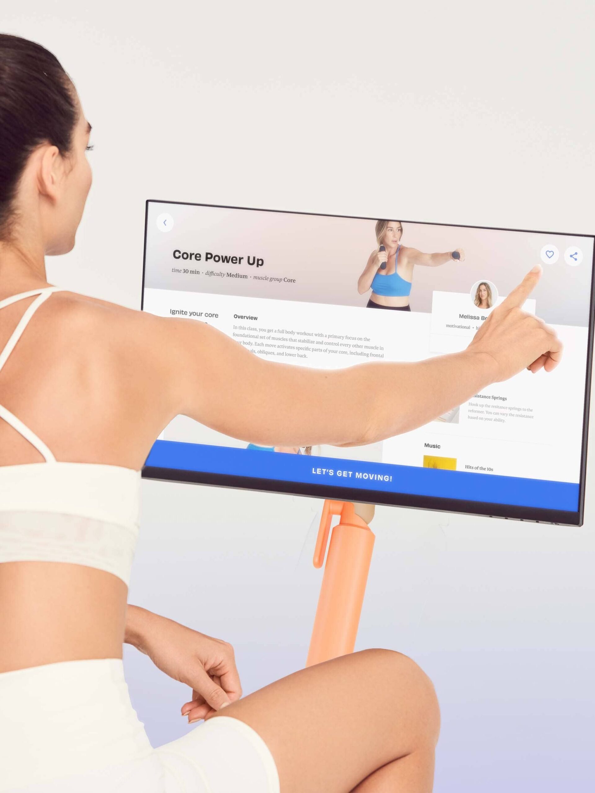 A woman in a white workout outfit interacts with a touchscreen displaying a fitness program titled "Core Power Up," while massage guns lay nearby, offering the perfect post-workout recovery.