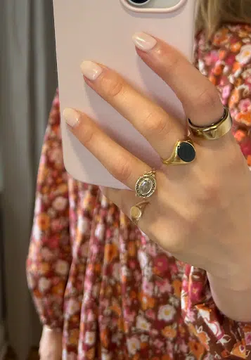 A hand with neatly manicured nails holds a pink phone, adorned with an Oura ring and three other stylish rings, against a floral-patterned fabric.