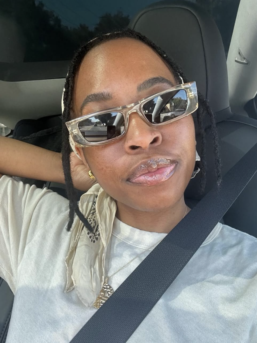 Person with sunglasses and a beige headscarf, seated in a vehicle, wearing a white shirt and seatbelt, looking at the camera.