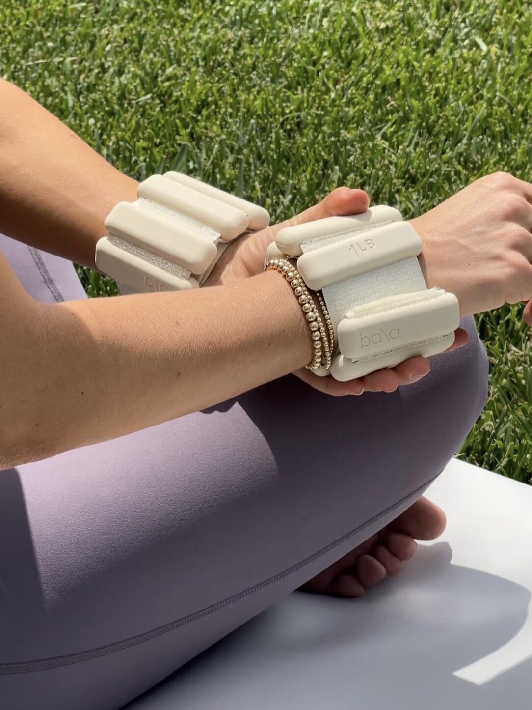 A person sits outdoors on a mat, wearing beige wrist weights and lavender leggings, with grass in the background. Nearby, a massage gun rests beside them, ready to enhance post-workout relaxation.