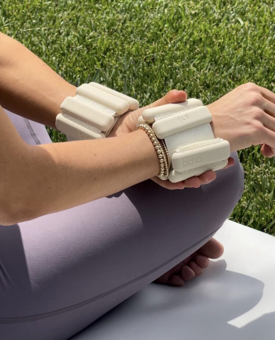 Person sitting on a yoga mat outdoors, wearing beige wrist weights and purple leggings, with a bracelet on one wrist. Nearby, a massage gun rests on the grass, ready to enhance post-yoga relaxation.